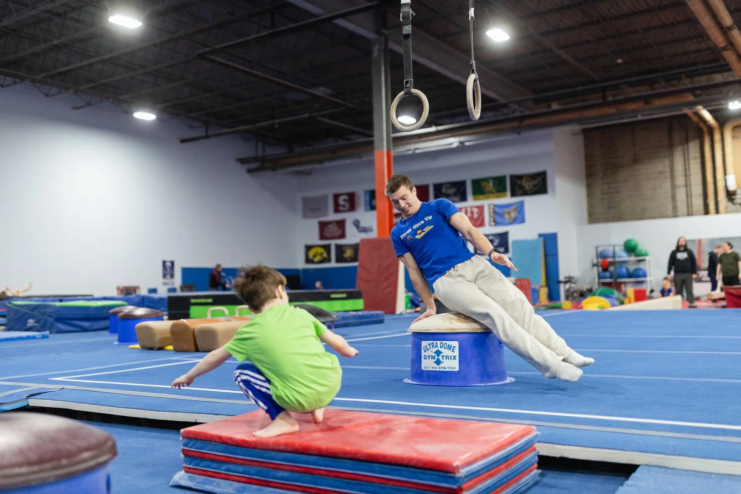 A gymnastics training facility with gymnastic rings hanging from the ceiling, blue padded flooring, and equipment like mats and vaulting tables. A coach is assisting a young child on a vaulting apparatus while another child is nearby. In the backgrou