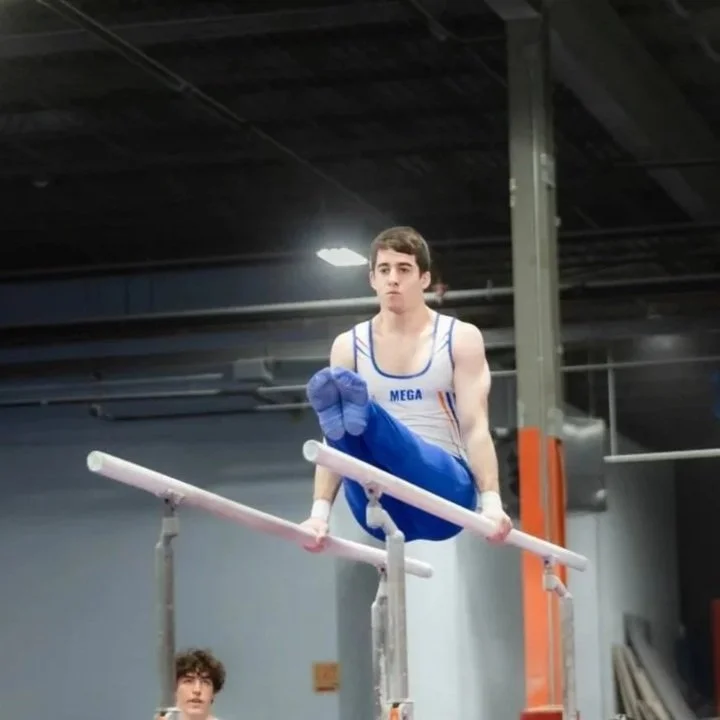 Young male gymnast in a white and blue uniform executing a vault move on parallel bars in an indoor gym, with a serious expression on his face.