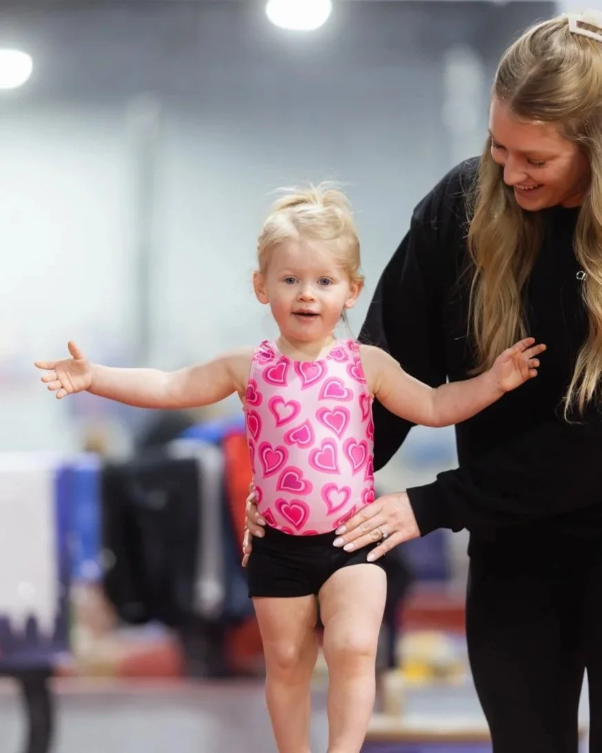Young girl with blonde hair in a pink leotard with heart patterns being supported by an adult woman with long blonde hair in a black outfit, in an indoor gym or sports facility.