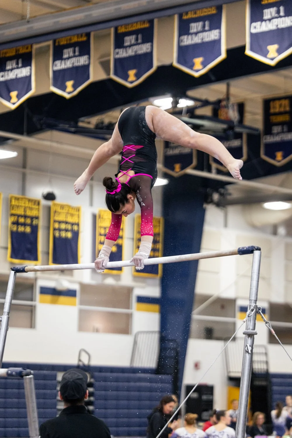 A female gymnast performs an upside-down move on the uneven bars during a competition in a gymnasium decorated with championship banners.