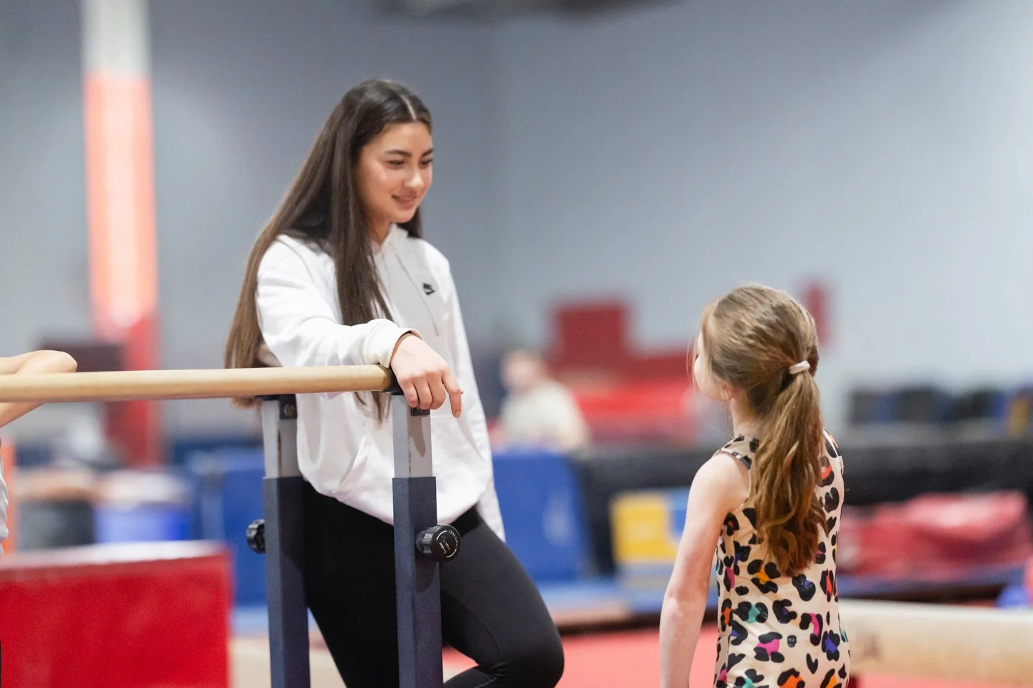 A young woman and a young girl conversing at a gymnastics facility, with the young woman leaning on a ballet bar and smiling.