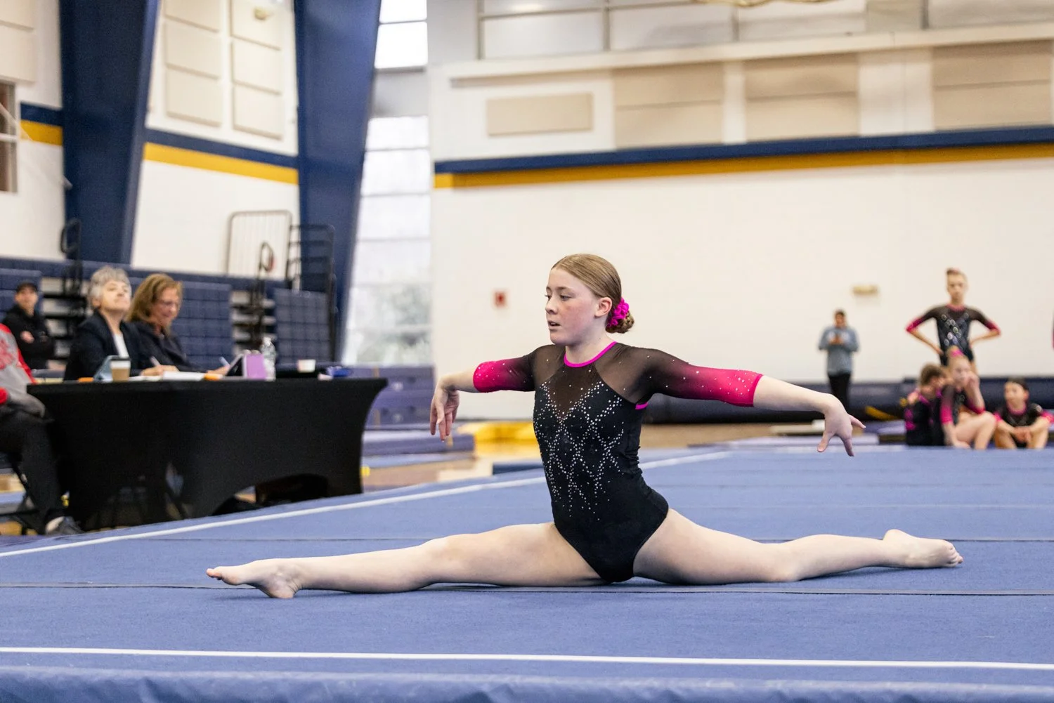A young gymnast in a black and pink leotard performs a split on the blue gymnastics floor during a competition, with judges watching on the side and other gymnasts in the background.
