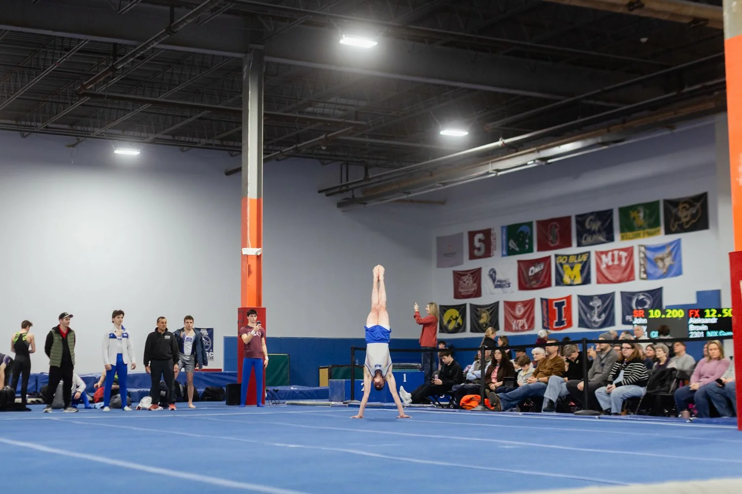 A gymnast is performing a handstand on a blue gymnasium floor while an audience watches. The background features banners and flags hanging on the wall.