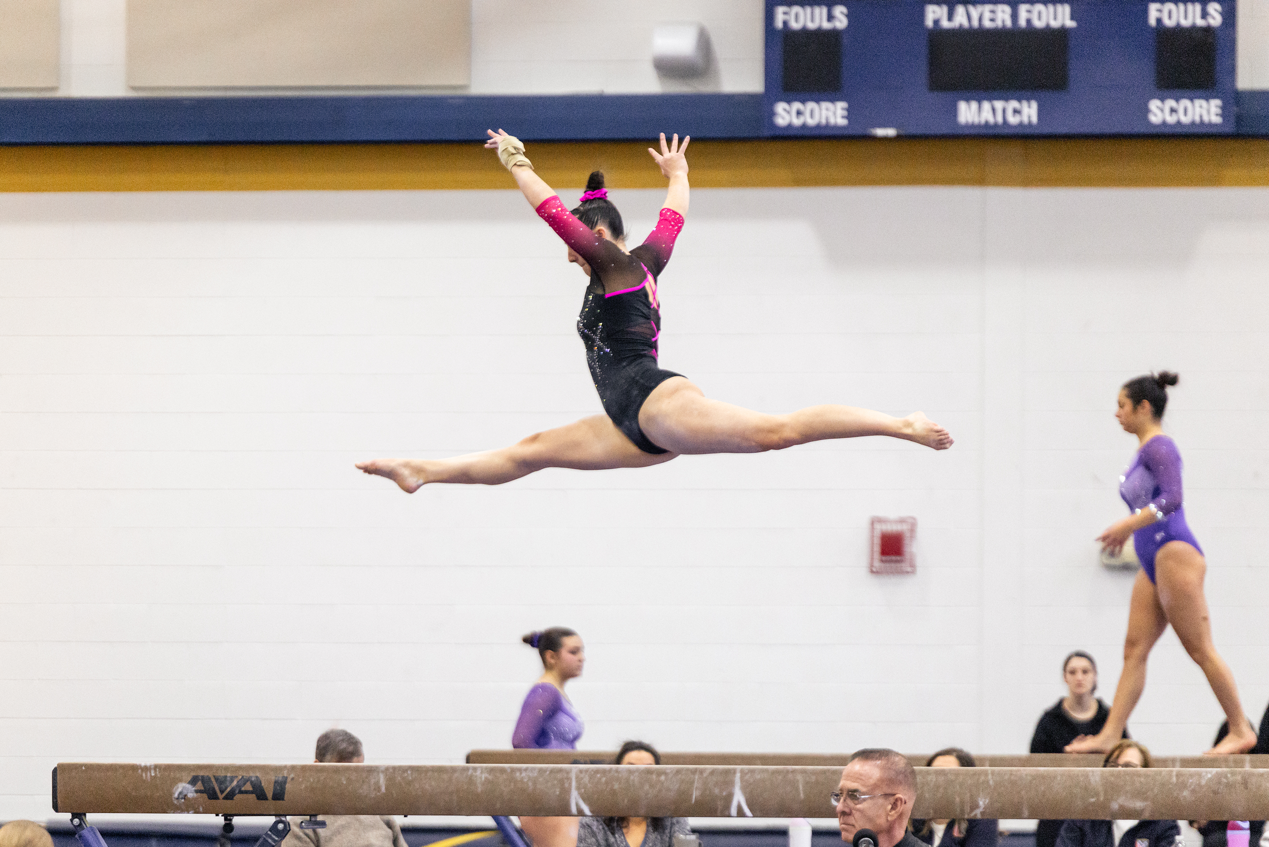 A female gymnast in a black and pink leotard performing a split leap on a balance beam during a gymnastics competition. In the background, other gymnasts and spectators are visible in an indoor gymnasium with a scoreboard displaying fouls and scores.