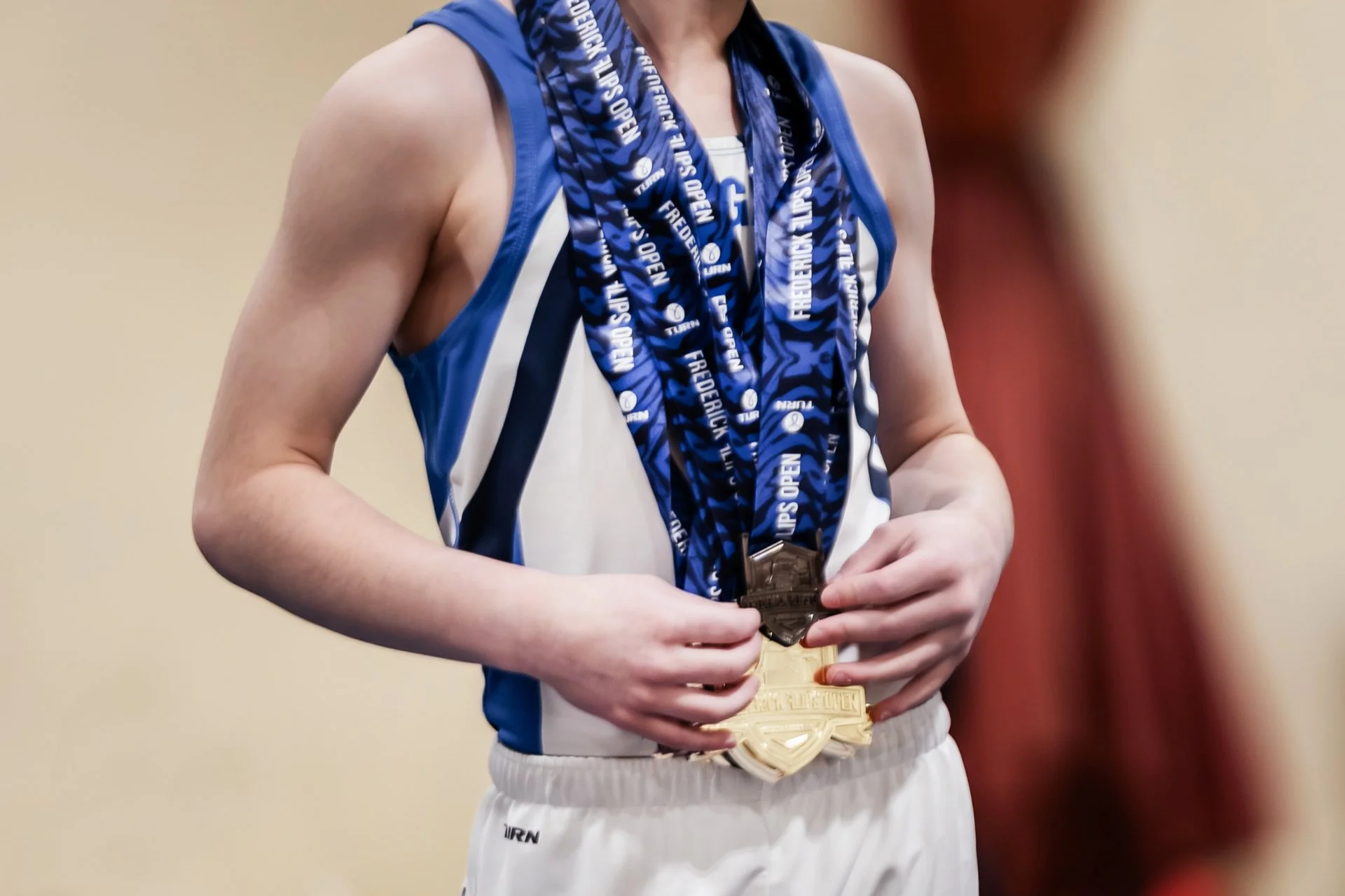 A person wearing a blue sports jersey and white shorts is holding multiple medals, with one medal hanging around their neck. The medals are gold, silver, and bronze, indicating first, second, and third place.