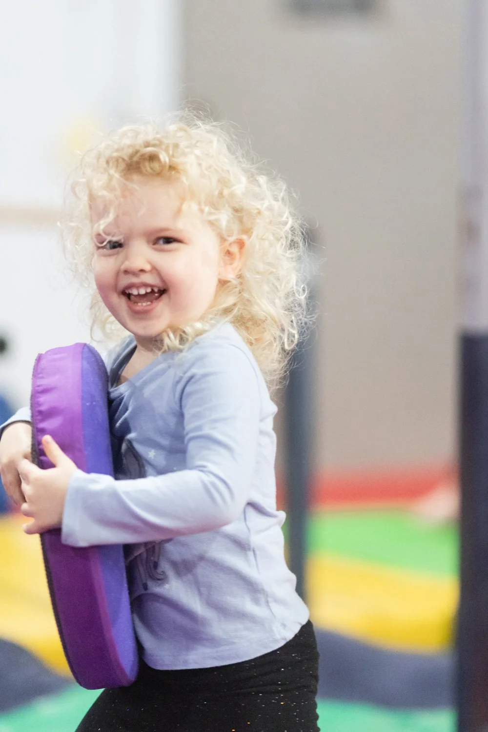 A young girl with curly blonde hair smiling and holding a purple object in an indoor play area.
