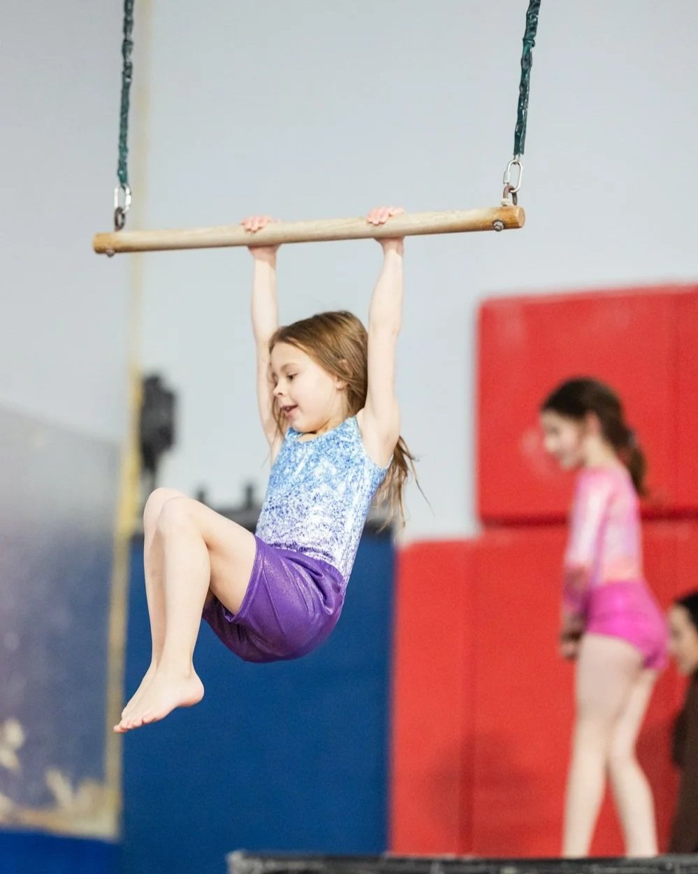 A young girl hanging from a gymnastics bar in a gym class, with other children blurred in the background.