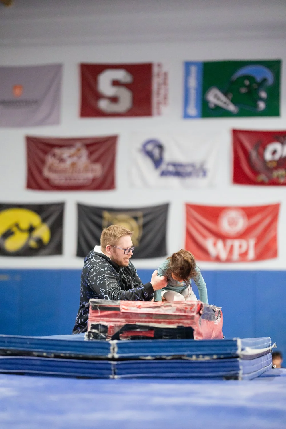 A man with glasses and a black hoodie helping a young girl with brown hair practicing gymnastics on a padded mat in an indoor gym, with various sports flags hanging in the background.