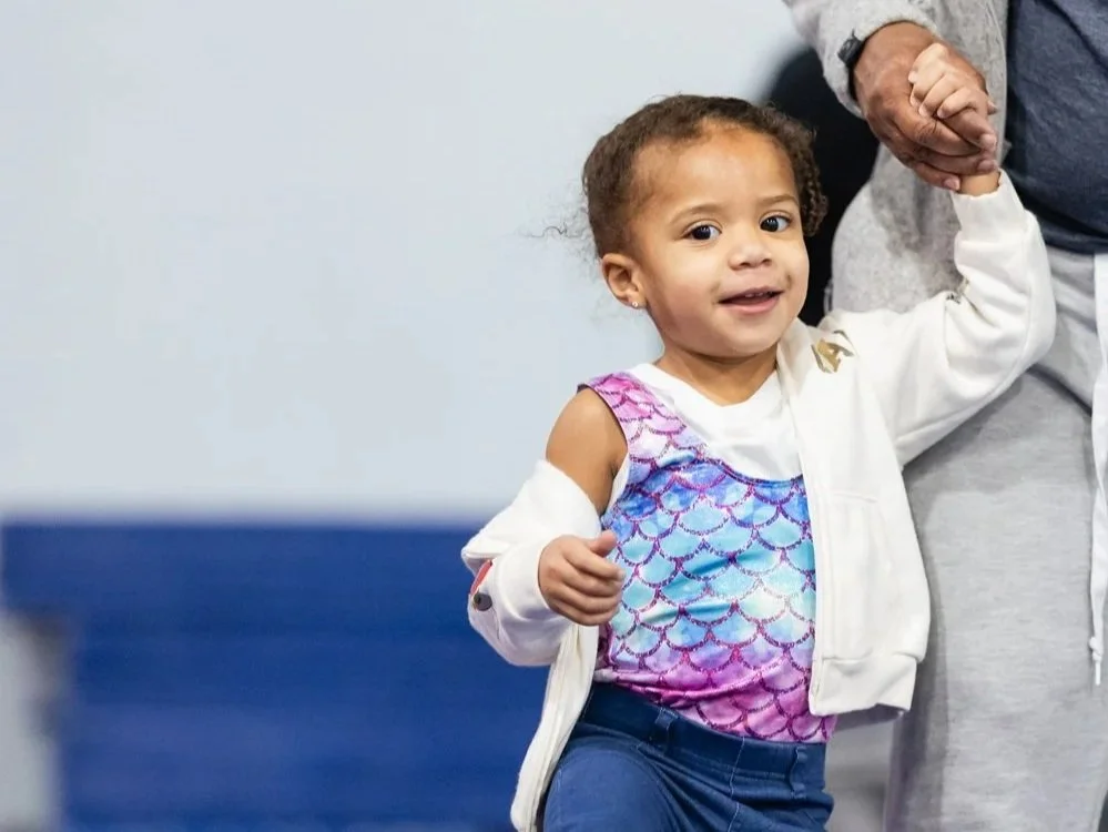 A young girl with curly hair in a white jacket receives support from an adult, holding her hand.