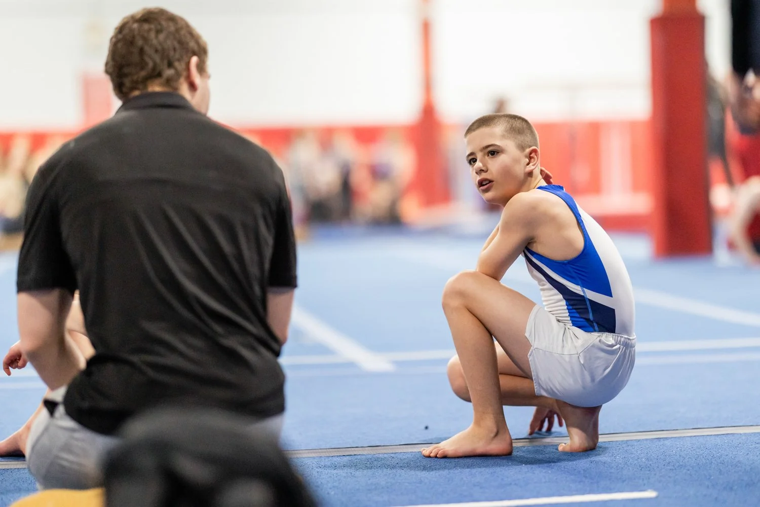 A young male gymnast in a blue and white singlet crouches on a blue gym mat, talking to a male coach who is sitting on the mat with his back to the camera, in an indoor gym.