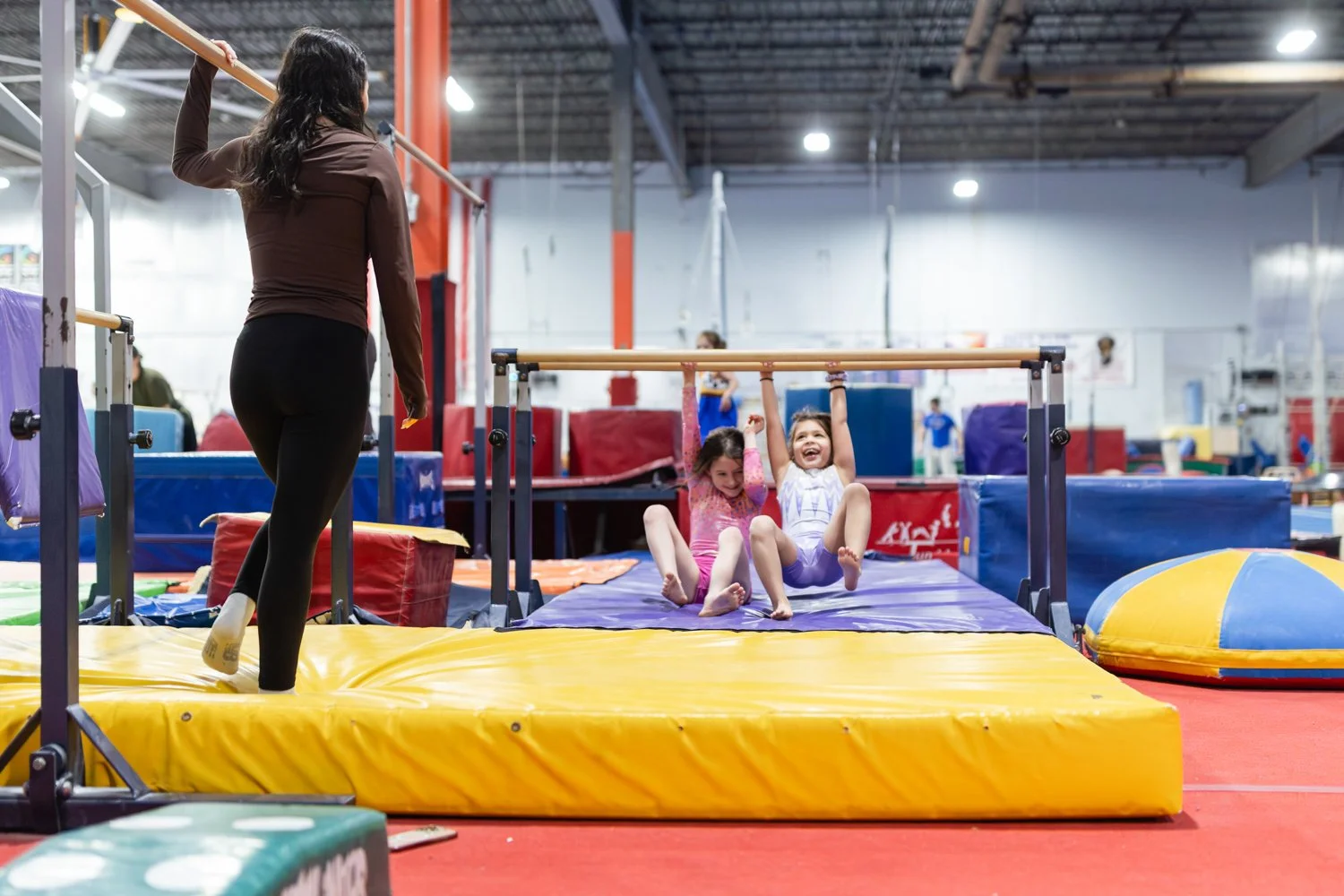 Two young girls sitting on a gymnastics mat, hanging from a horizontal bar, with a woman standing nearby in an indoor gymnastics facility.