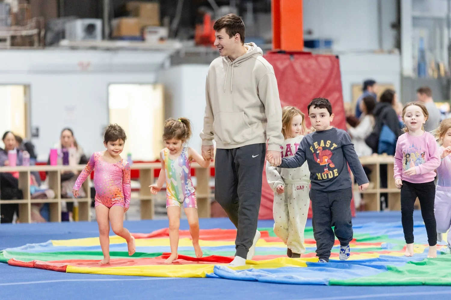 Young children in colorful clothes walking on a rainbow-colored mat in an indoor gym, supervised by an adult man.