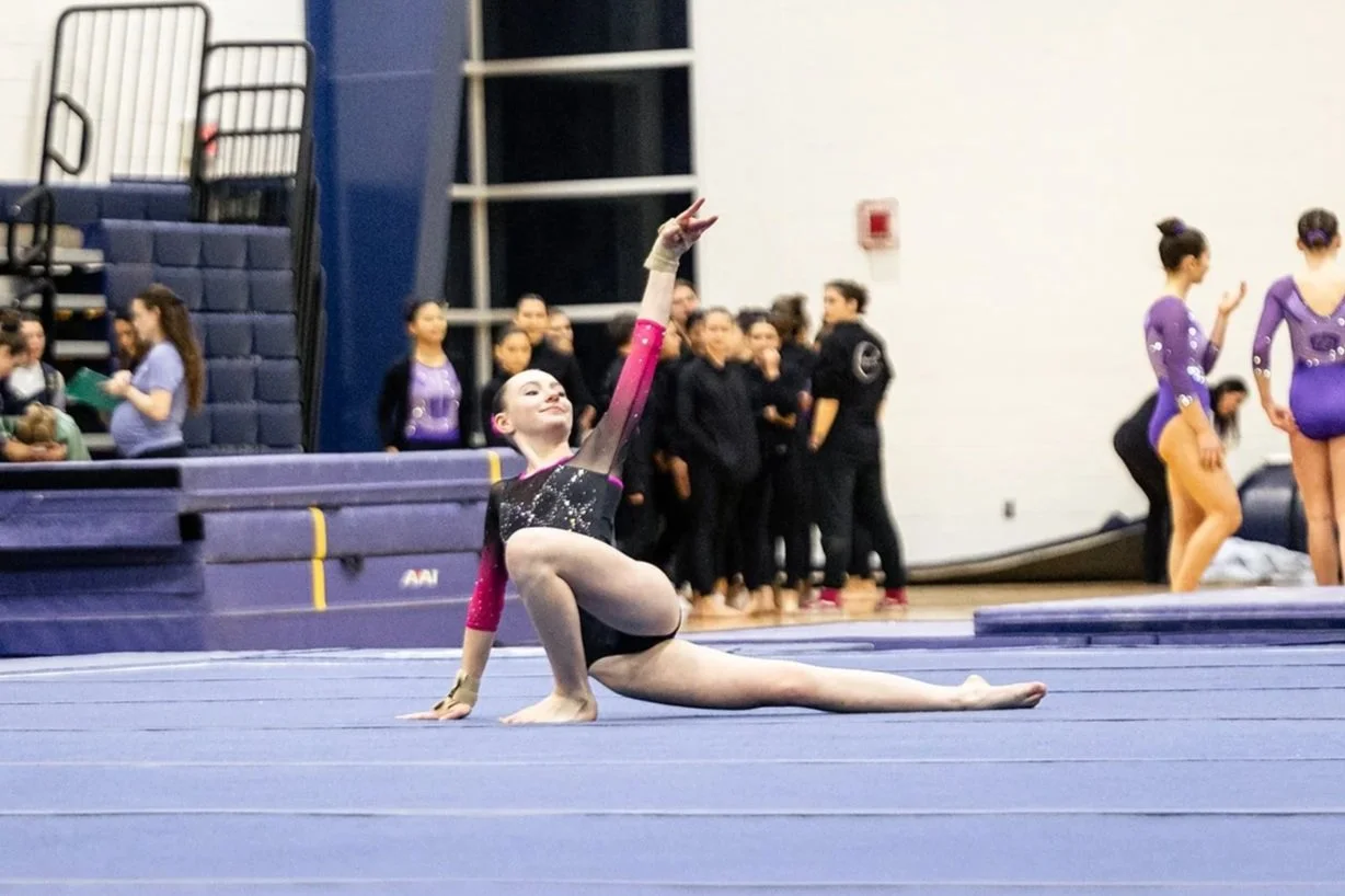 A gymnast performs a routine on a blue gymnastics mat, extending one arm upwards and one leg out to the side in a pose during a competition. In the background, there are other gymnasts and coaches, some in purple leotards, and spectators sitting in the bleachers.