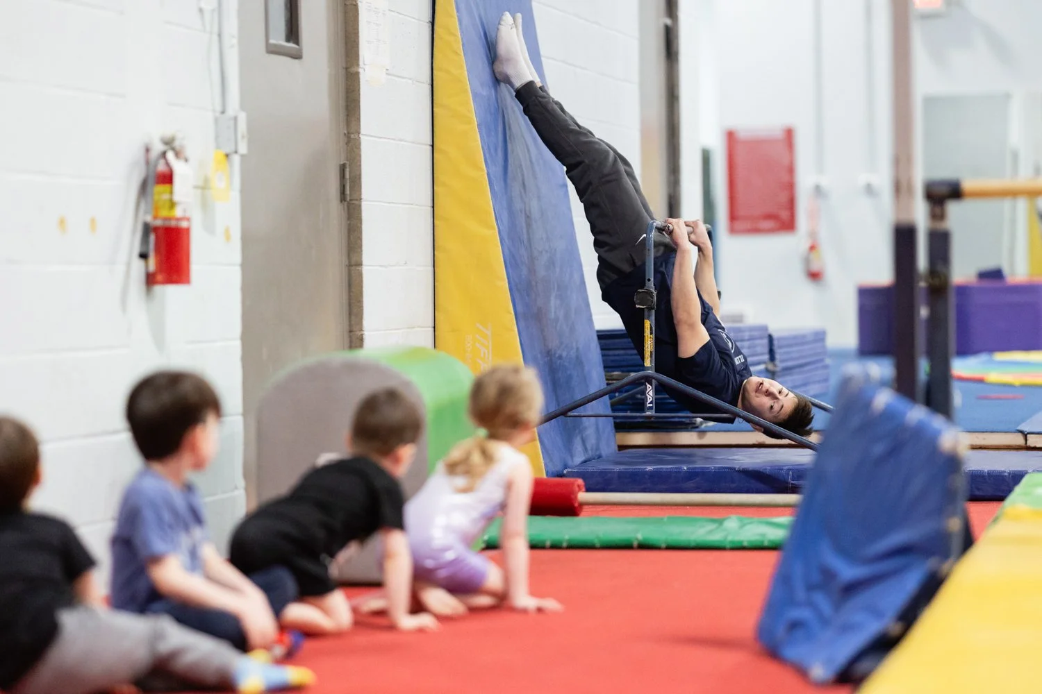 A man hanging upside down on a trampoline in a gymnastics gym, with children kneeling on the mat watching him.