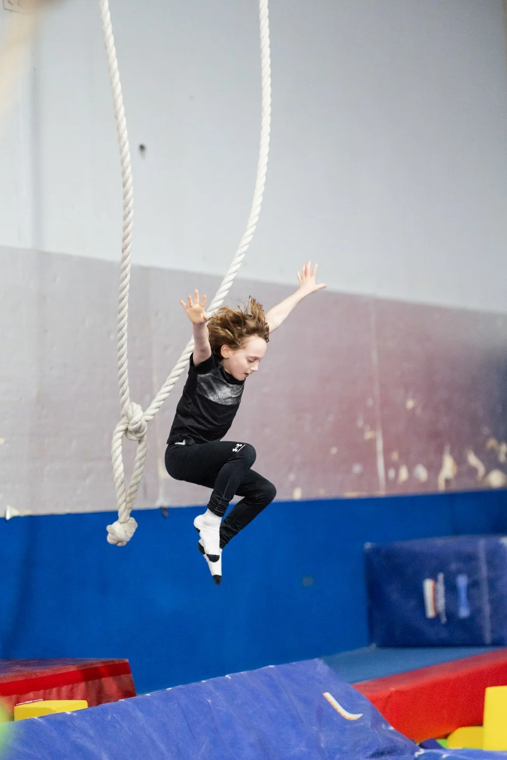 Young girl in black athletic clothing and socks jumps on gymnastics foam pit at indoor gym.