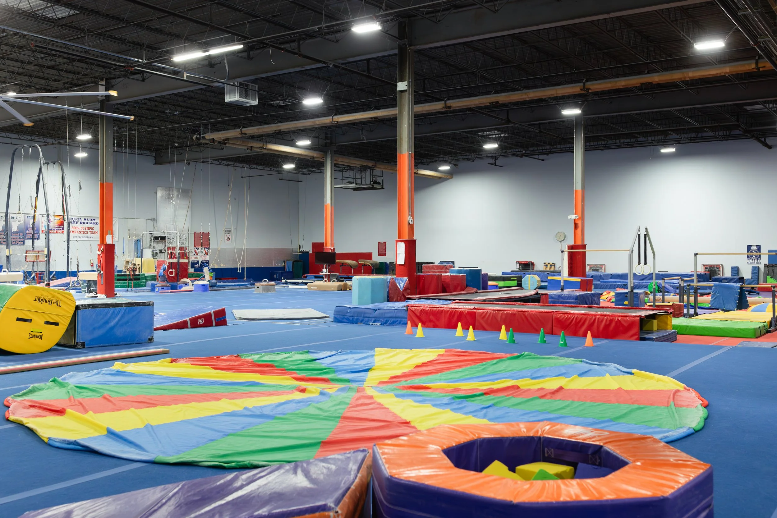 An indoor gymnastics training facility with various equipment including mats, trampolines, and climbing apparatus, under a high ceiling with industrial lighting.