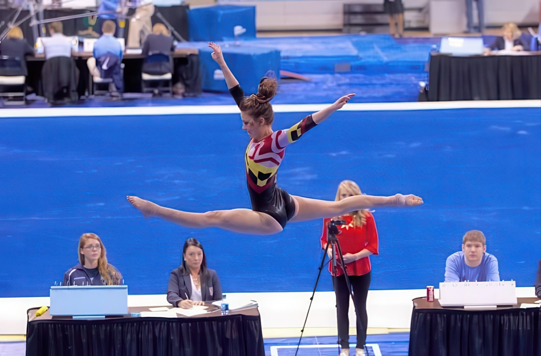 A female gymnast performing a split leap in mid-air during a gymnastics competition, with judges and officials seated at tables below on a blue mat.