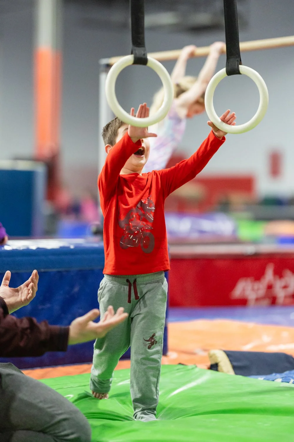 Young boy in red shirt and gray sweatpants is playing on gymnastic rings, reaching up to grab them, with an instructor or friend nearby clapping and encouraging him.