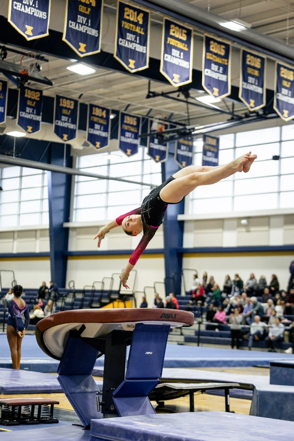 A young gymnast performing a vault during a gymnastics competition, with a crowd seated in the background and banners hanging from the ceiling celebrating past championship wins.