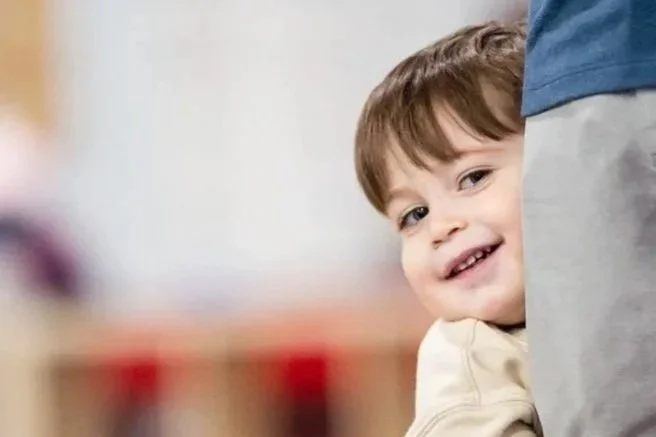 A young boy smiling, peeking out from behind an adult, in an indoor setting.