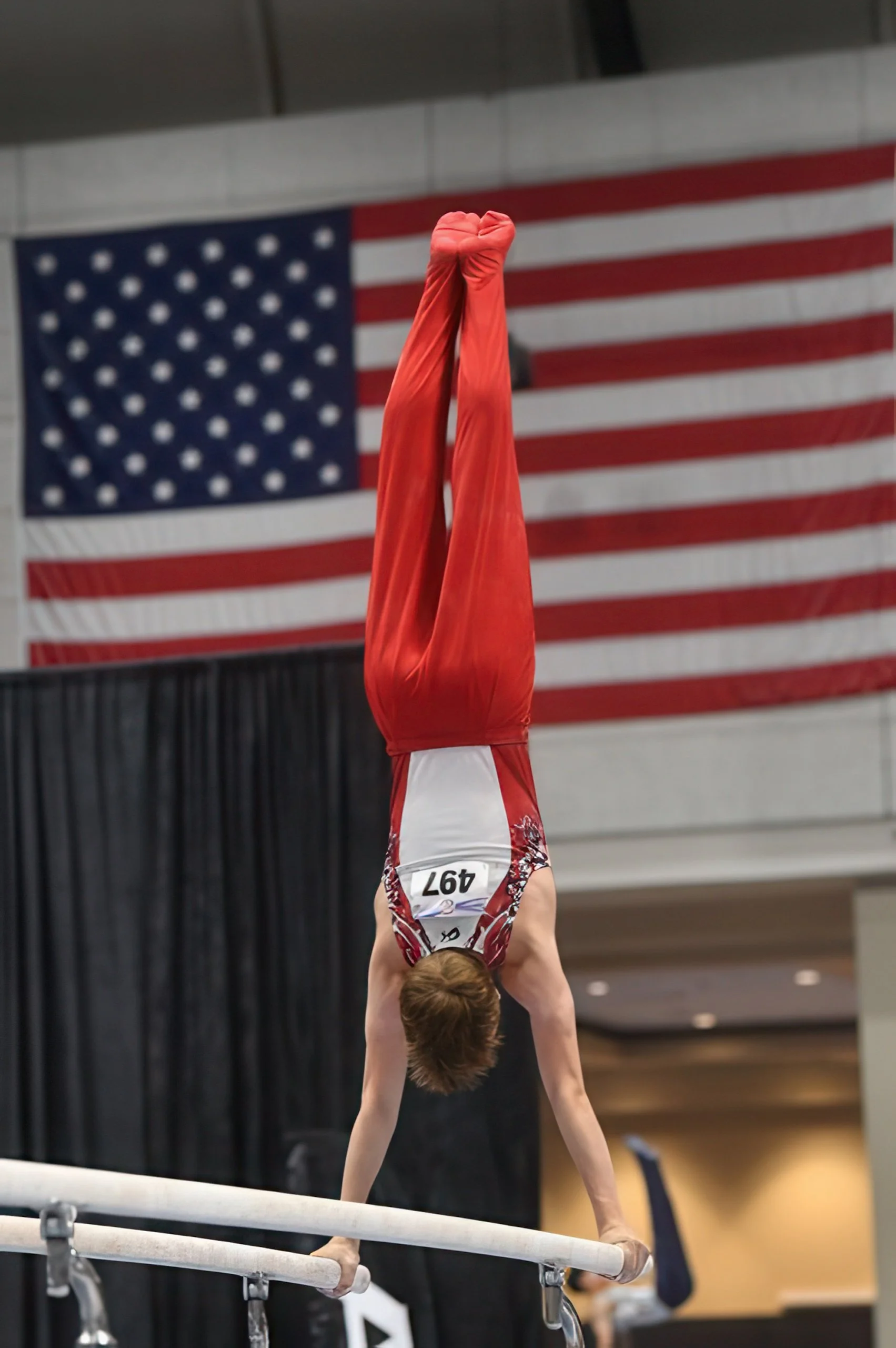 A gymnast performing a handstand on the balance beam with an American flag hanging in the background.