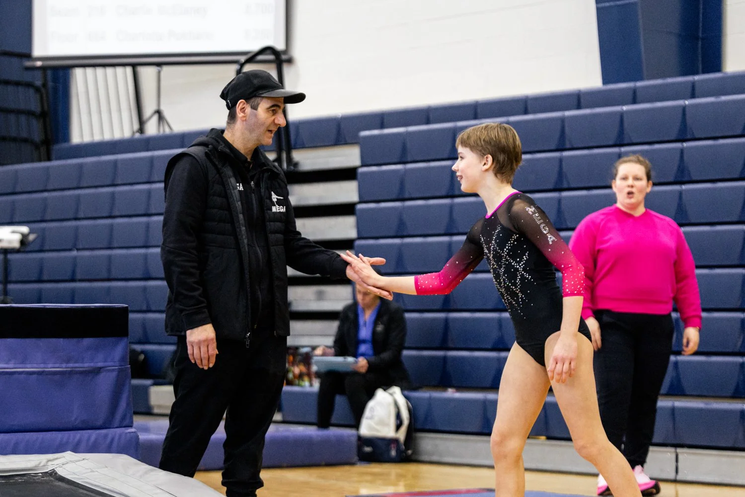 A young female gymnast in a black and pink leotard shaking hands with a coach or judge in a gymnasium. A woman in a pink top stands in the background.
