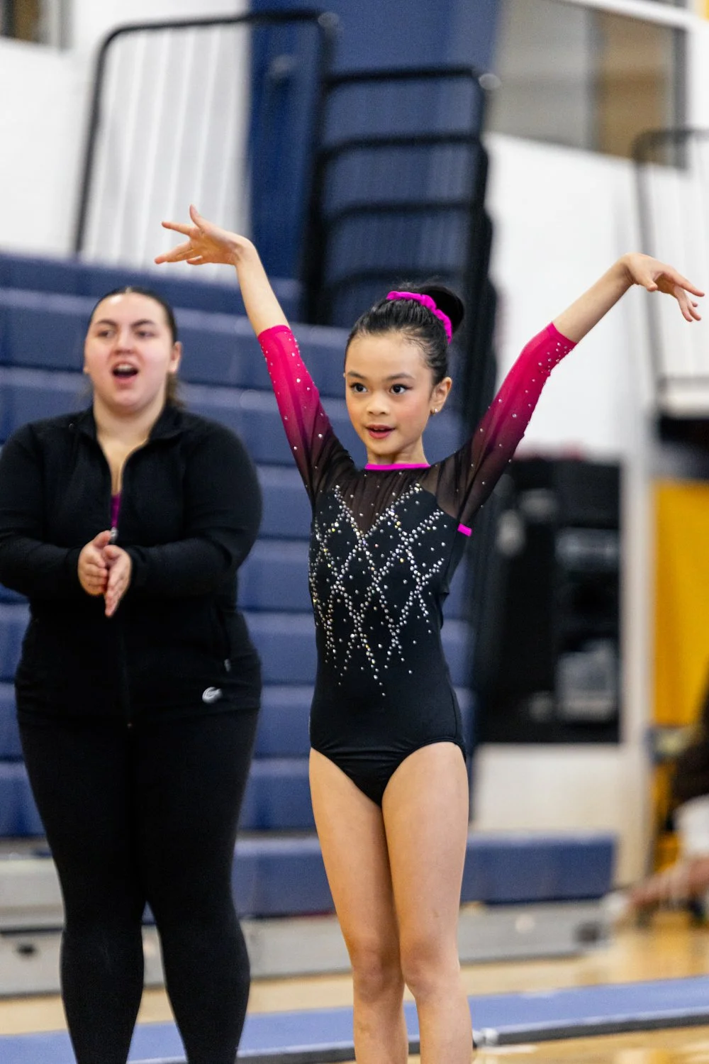 A young girl in a black gymnastics leotard with pink accents and rhinestone designs, performing a routine with her arms raised, while an instructor or coach stands behind her clapping in a gymnasium.