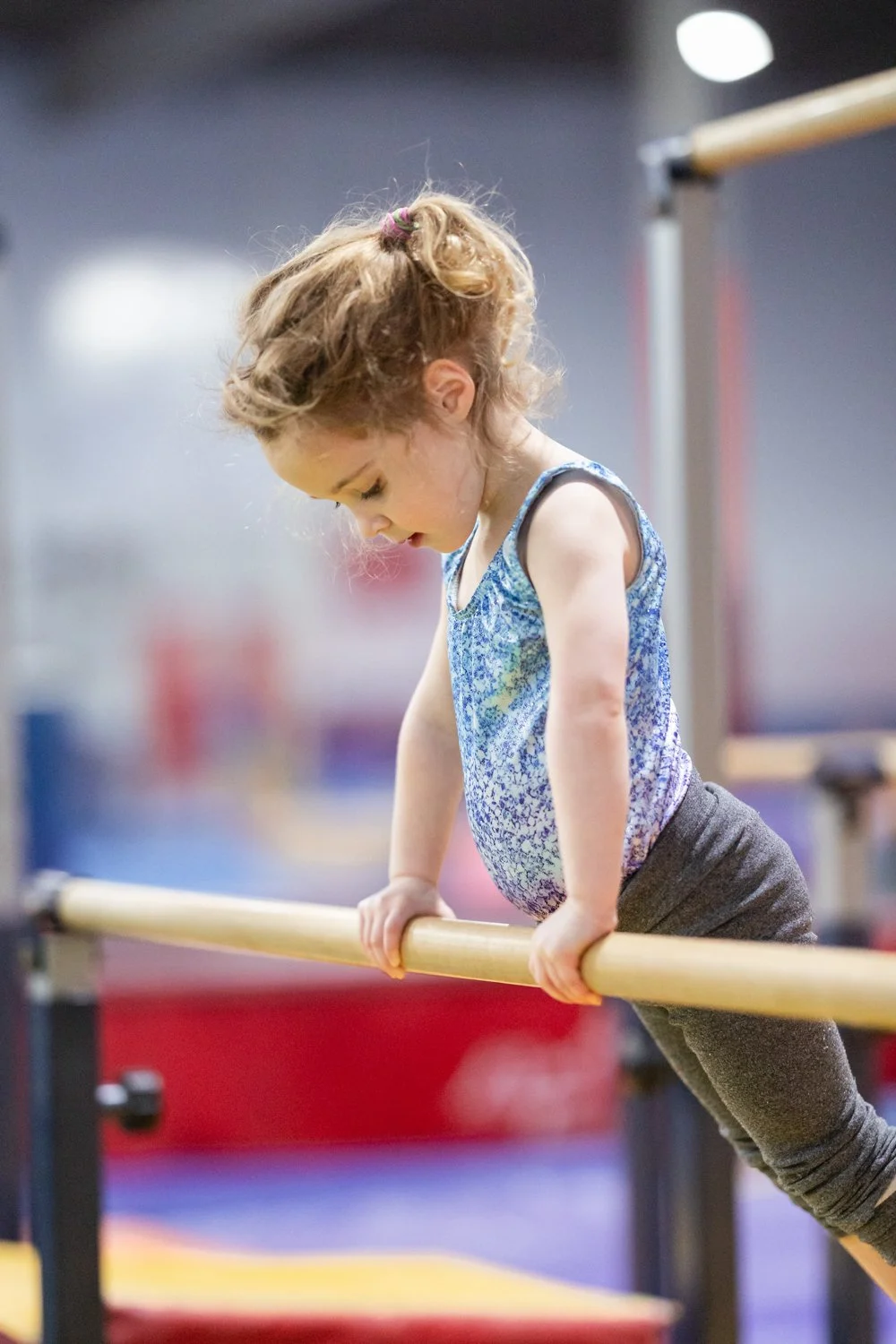 A young girl with curly hair doing a fitness move on a horizontal bar at a gym or gymnastics facility.
