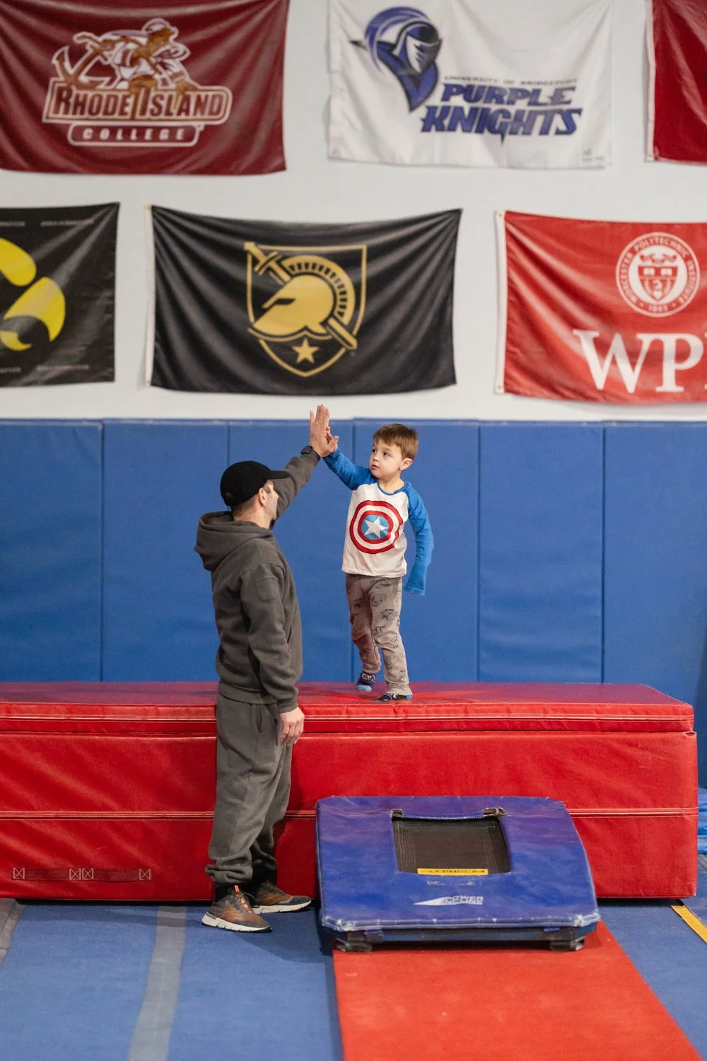 A young boy in a Captain America shirt giving a high five to an adult man in a hoodie in a gymnasium with banners on the wall.