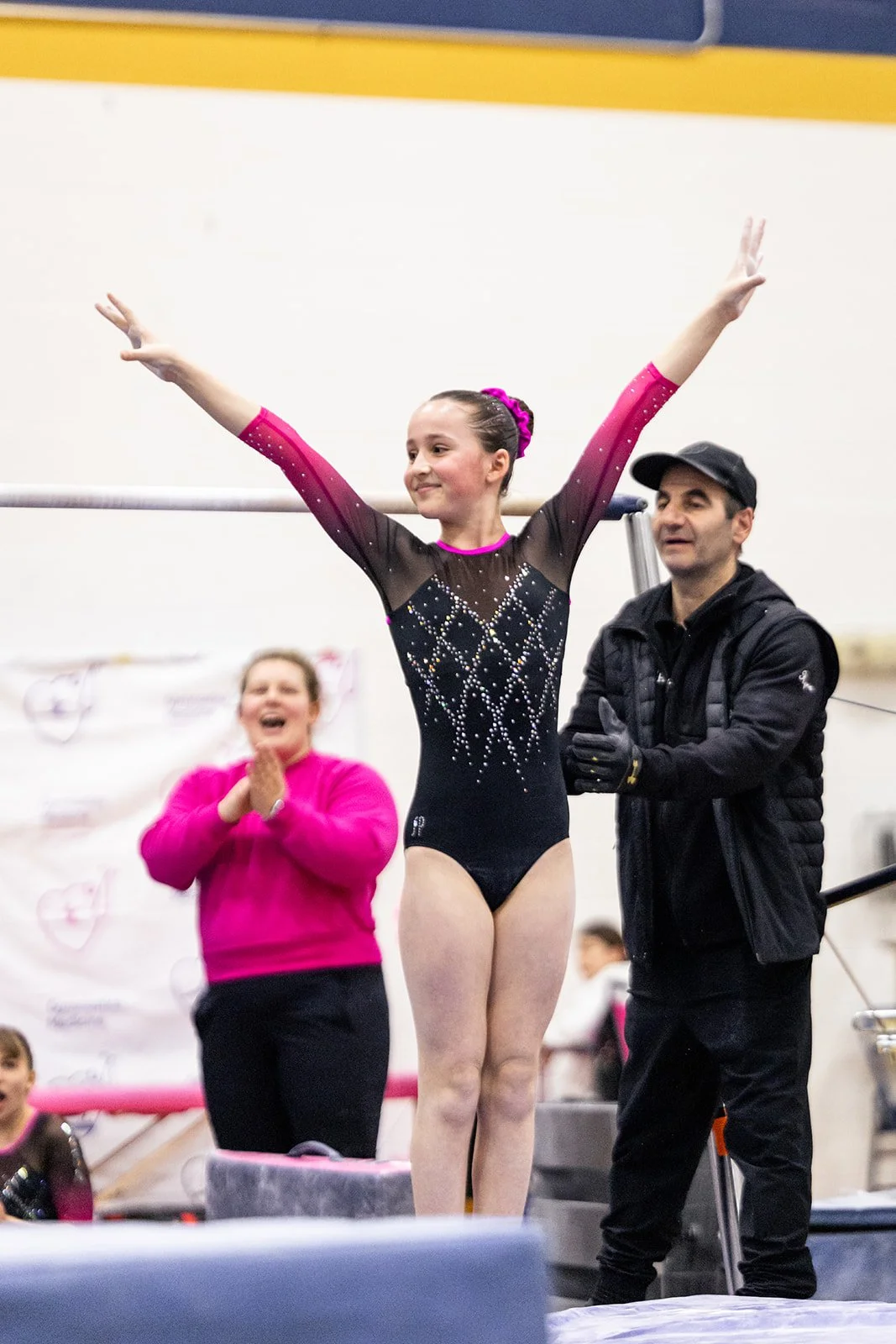 A young female gymnast stands on a balance beam with her arms raised in celebration, wearing a black and pink leotard decorated with rhinestones. An adult coach or judge stands beside her, and another woman in a pink shirt claps and smiles in the bac