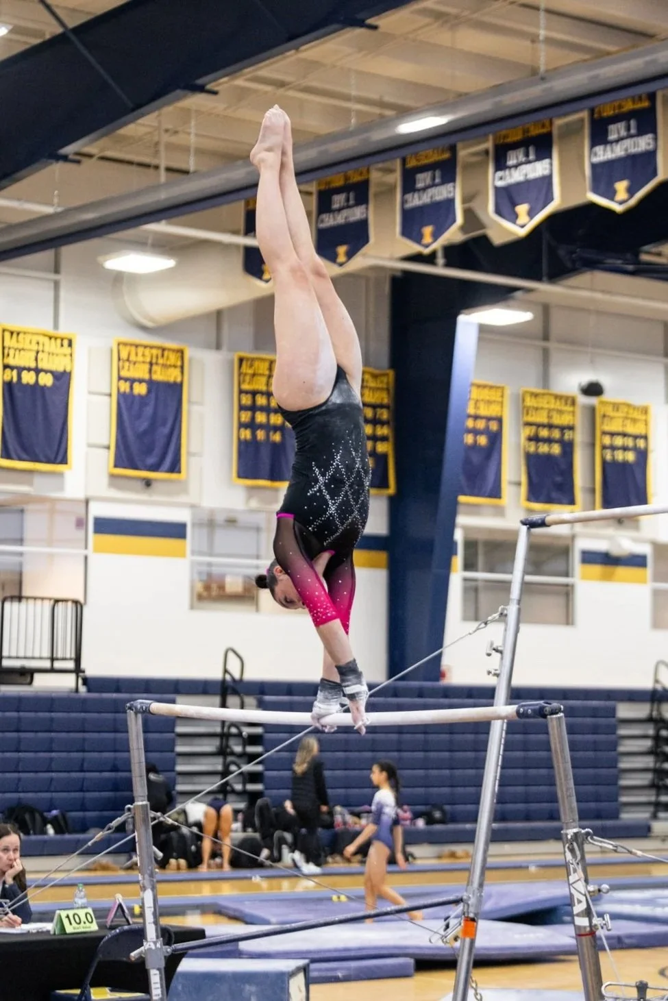 A female gymnast performs a handstand on the uneven bars in a gymnasium with banners hanging from the ceiling and people in the background.