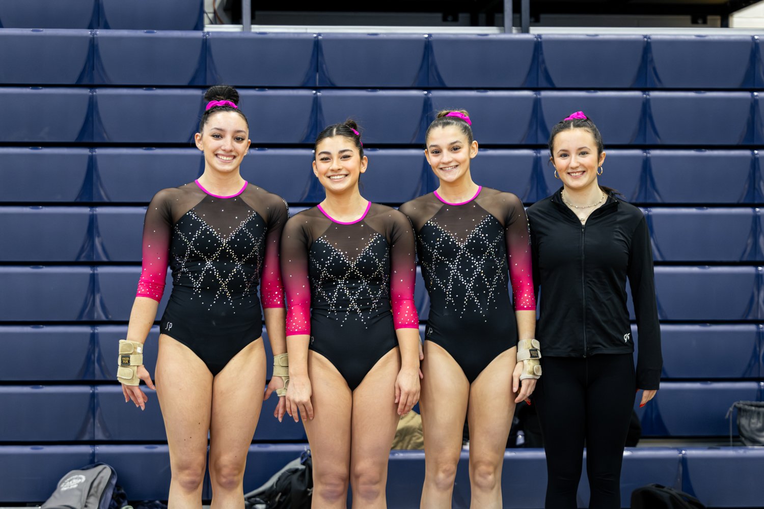 Four female gymnasts standing side-by-side in front of blue bleachers, wearing matching black and pink leotards with rhinestone designs, with the fourth woman dressed in black athletic wear.