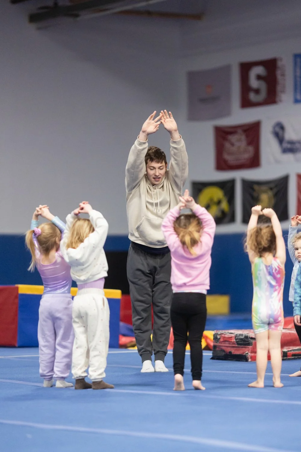 A young female instructor leading young girls in a gymnastics class, demonstrating a stretching exercise in an indoor gymnasium with banners hanging on the wall.