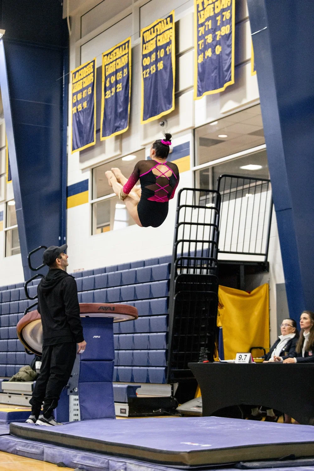 A gymnast in a black and pink leotard performing a vault in an indoor gymnasium, with judges seated at a table nearby and banners hanging on the wall.