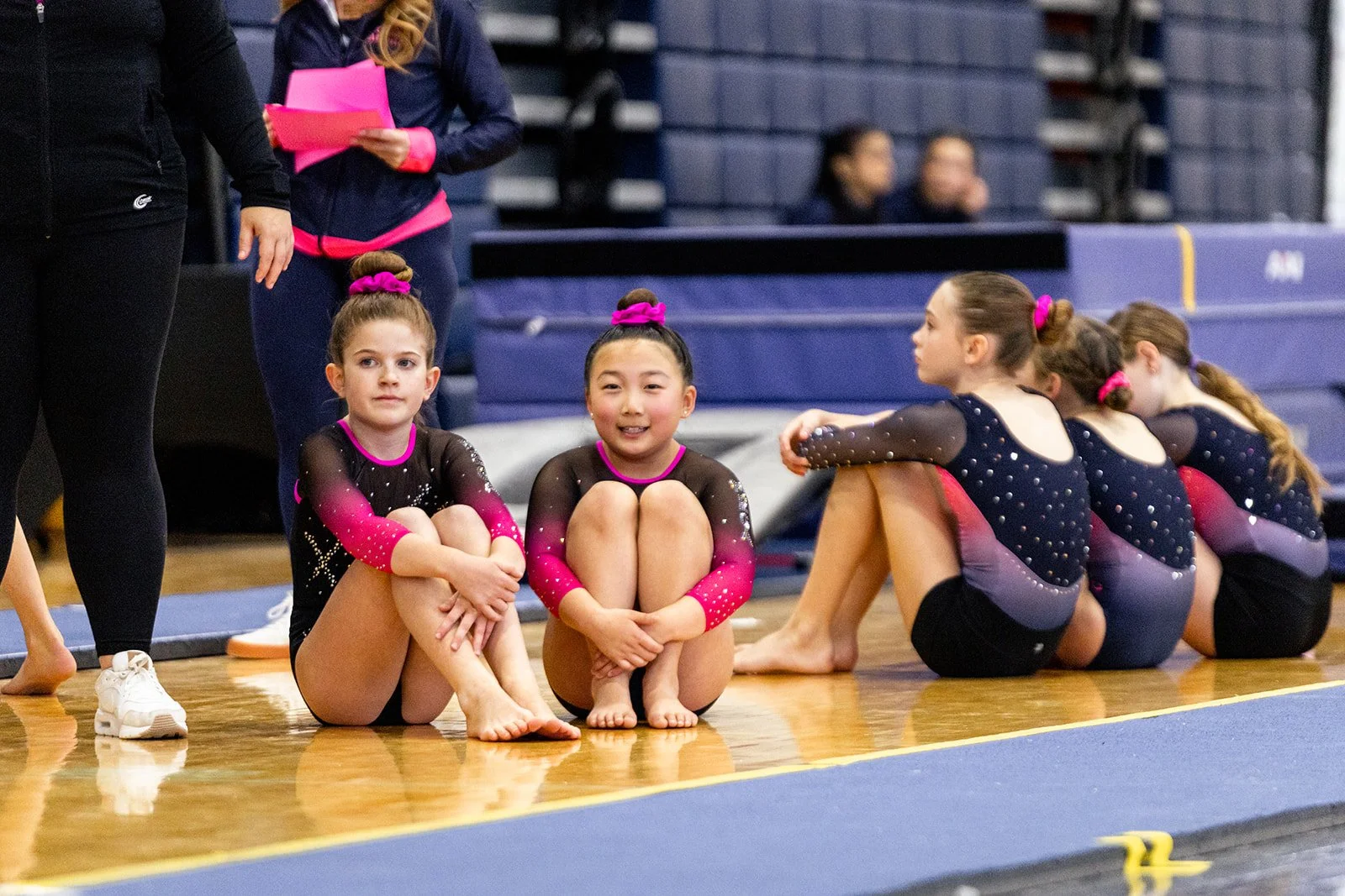 Group of young female gymnasts with matching black and pink leotards sitting on the floor of an indoor gymnasium, some sitting cross-legged, some sitting with knees up, with gymnastics mats in the background.