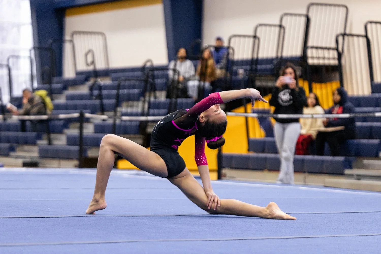 A young female gymnast is performing a split on the gymnasium floor during a competition. She is wearing a black and pink leotard with sparkles and has her hair tied in a bun with a pink ribbon. In the background, there are spectators sitting on blea