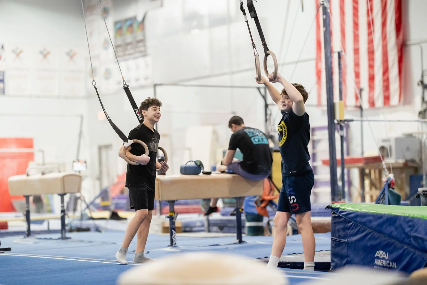 Two boys at an indoor gymnastics facility using gymnastic rings, smiling and engaging in training, with a coach or trainer in background.