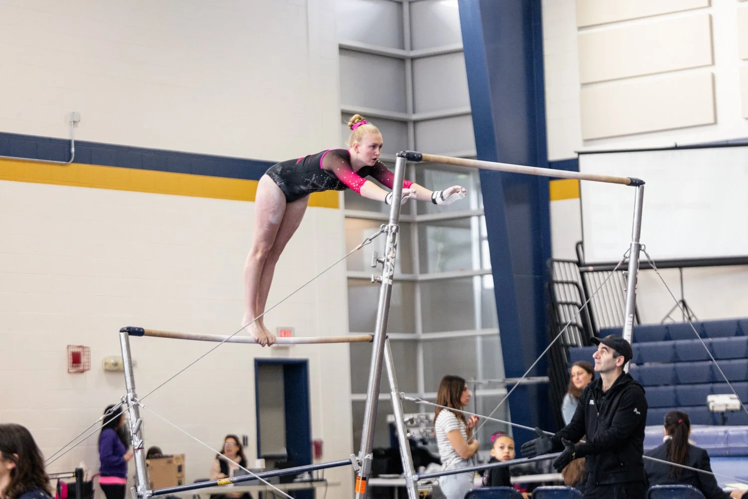 A young female gymnast practicing on the uneven bars in a gymnasium with spectators and coaches watching.