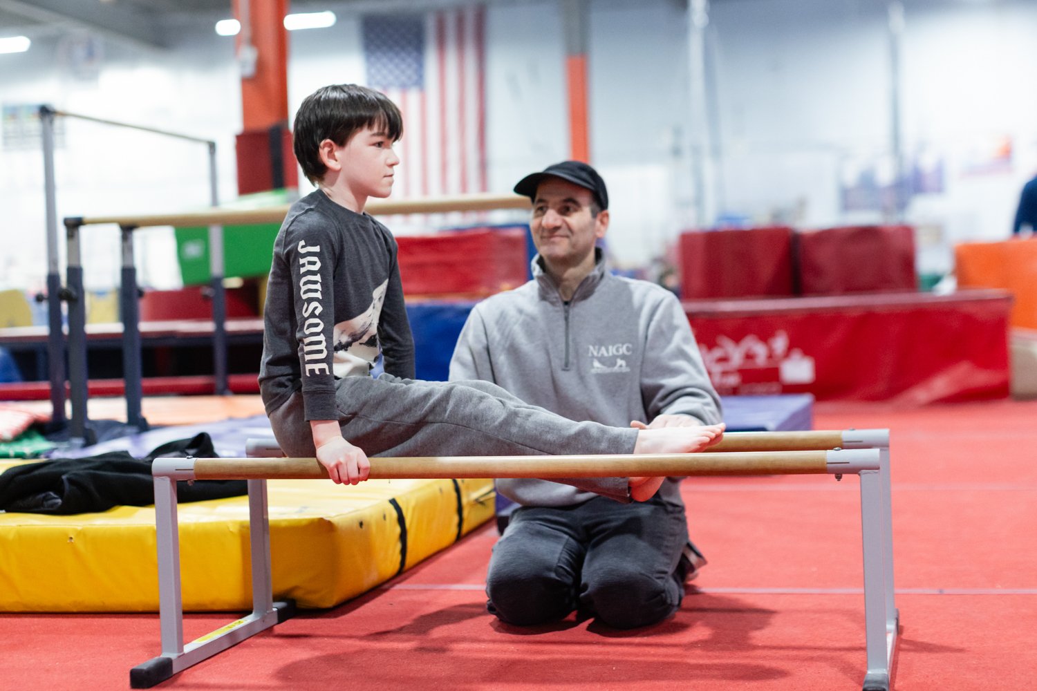 A young boy sitting on a balance beam with a coach kneeling beside him, indoors at a gymnastics facility.