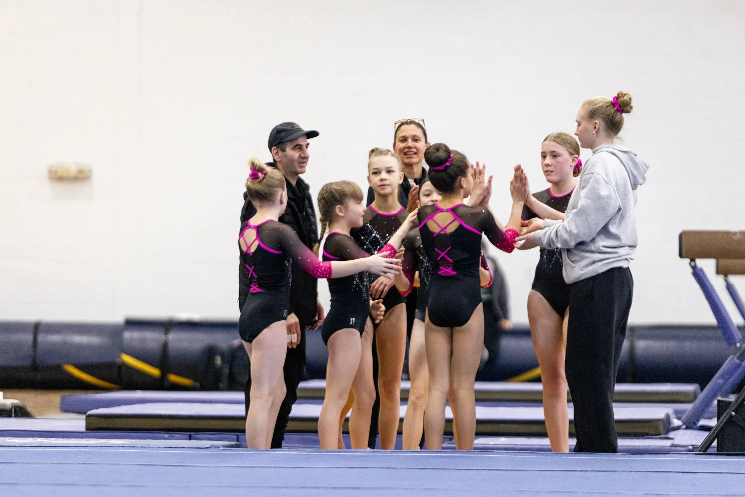 A group of young female gymnasts in black and pink leotards with matching hair accessories show high-fives and smiles as their coach and another adult give them encouragement in a gymnasium.