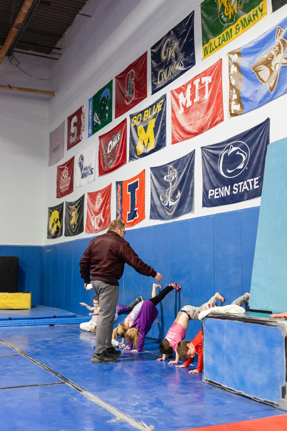 Kids doing handstands on a gym floor while a coach supervises, with sports banners hanging on the wall.
