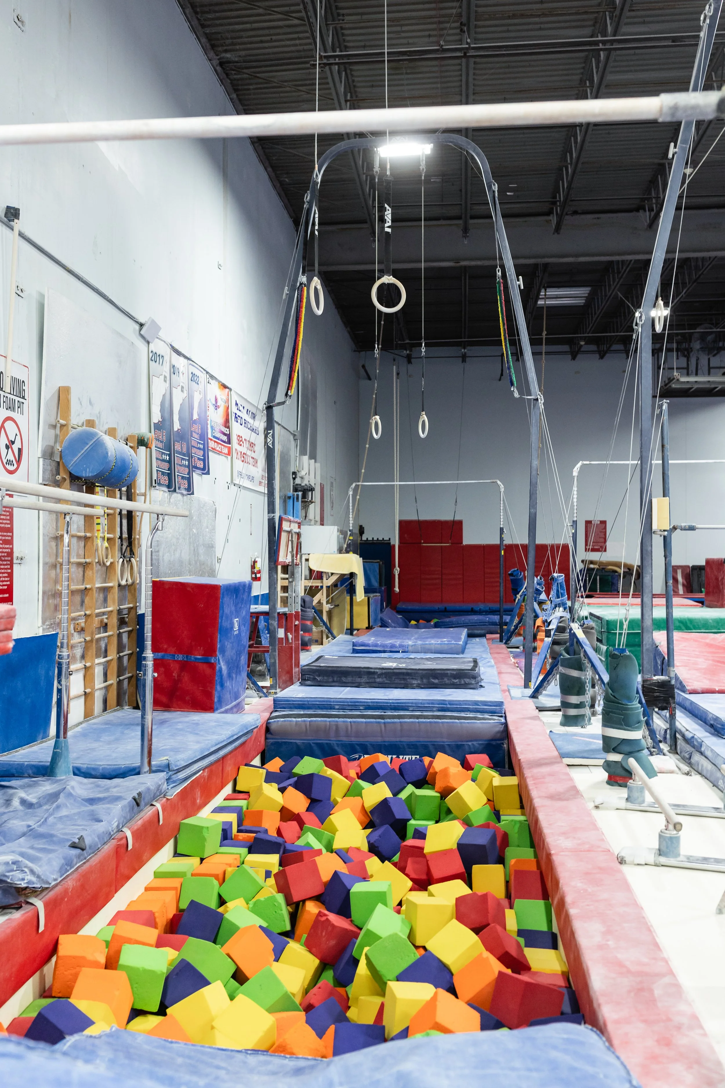 Indoor gymnastics training area with colorful foam blocks, padded mats, gymnastic rings, bars, and climbing equipment.