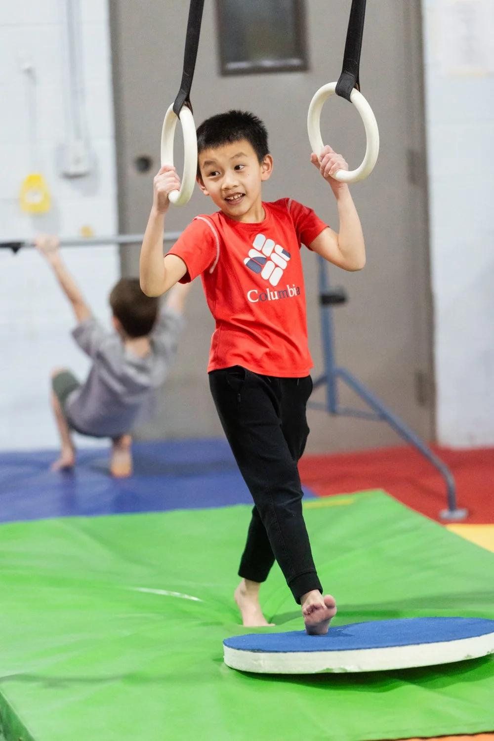A young boy in a red Columbia t-shirt balancing on a round platform while holding gymnastic rings in an indoor gym. Another child is in the background doing a handstand.