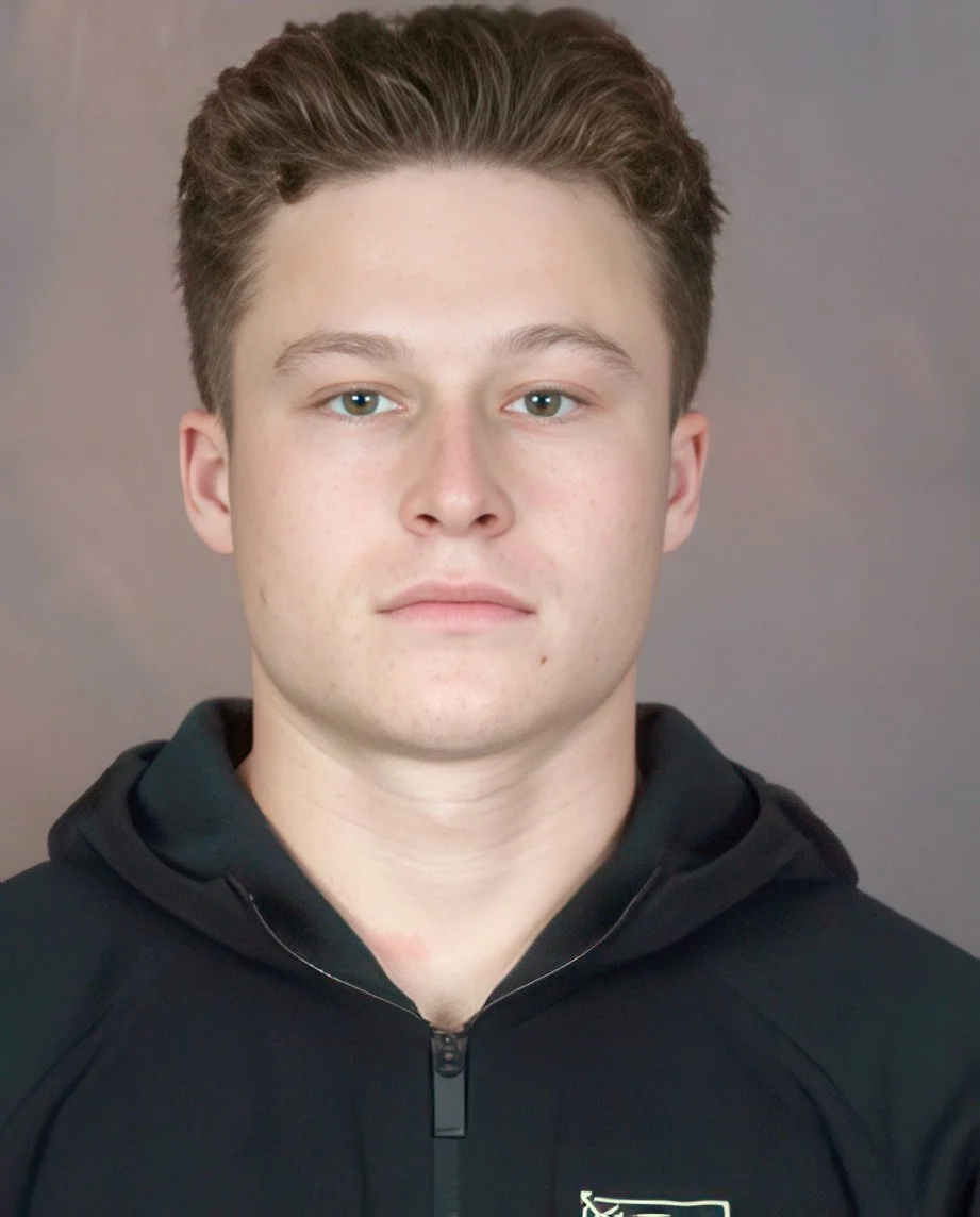 Close-up of a young man with light skin, brown hair, wearing a black zip-up hoodie with a small logo on the chest, neutral expression, against a plain background.