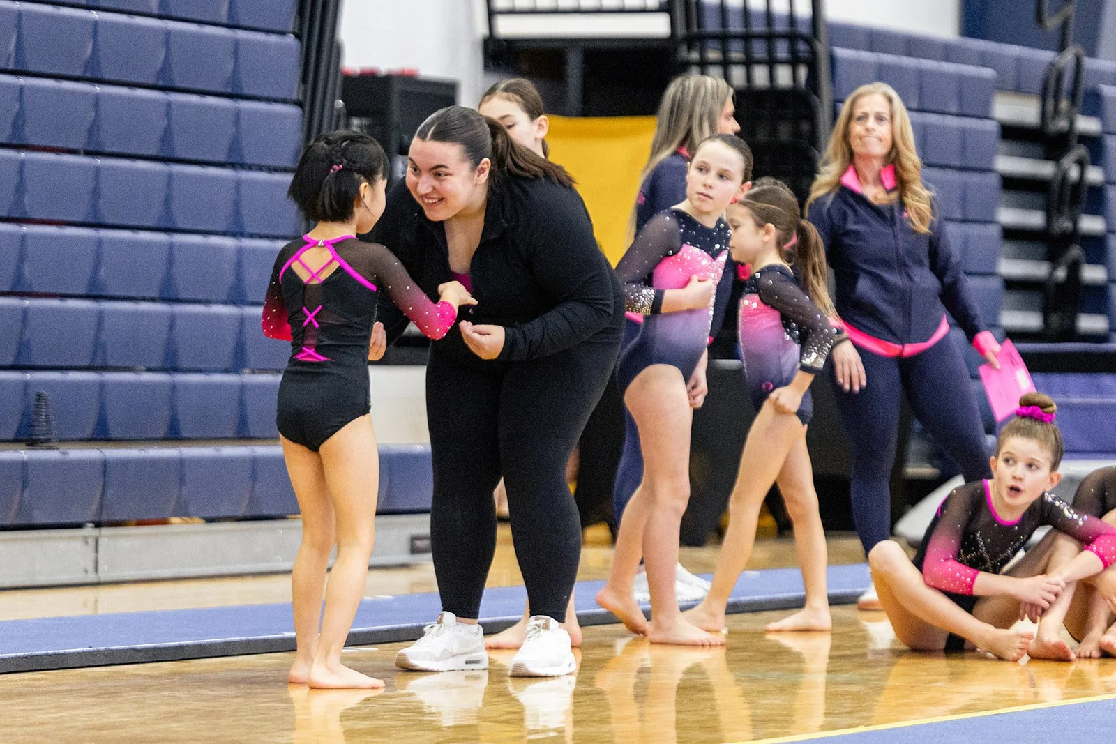 A coach is talking to a young gymnast in a black and pink leotard, with other young gymnasts and a woman with blonde hair observing in the background, inside a gymnasium.