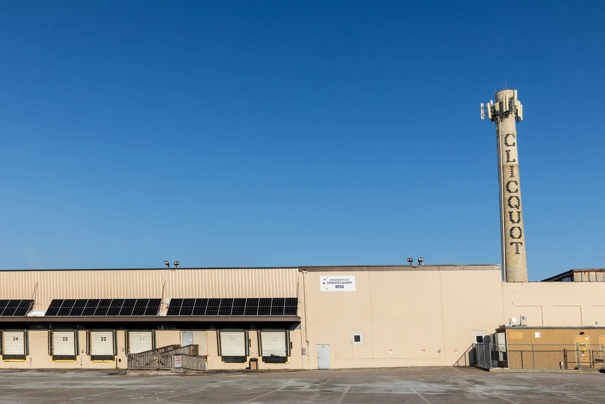 Large beige building with solar panels on the roof, a sign reading 'Massachusetts Elite Gymnastics Academy', and a tall tower with multiple antennas and the word 'Chicopee' displayed vertically, under a clear blue sky.