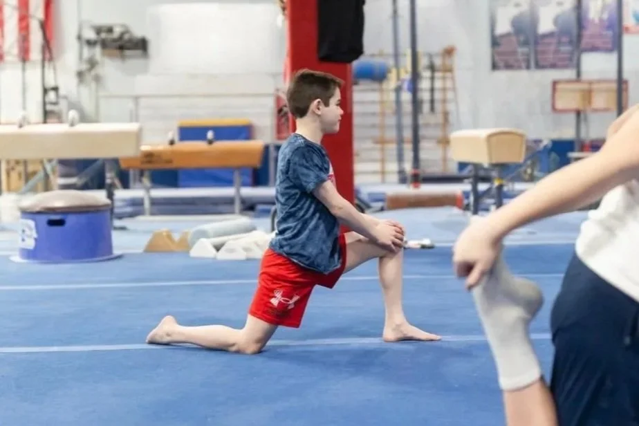A young boy practicing gymnastics in a gymnasium, wearing a blue shirt and red shorts, lunging on the blue mat.