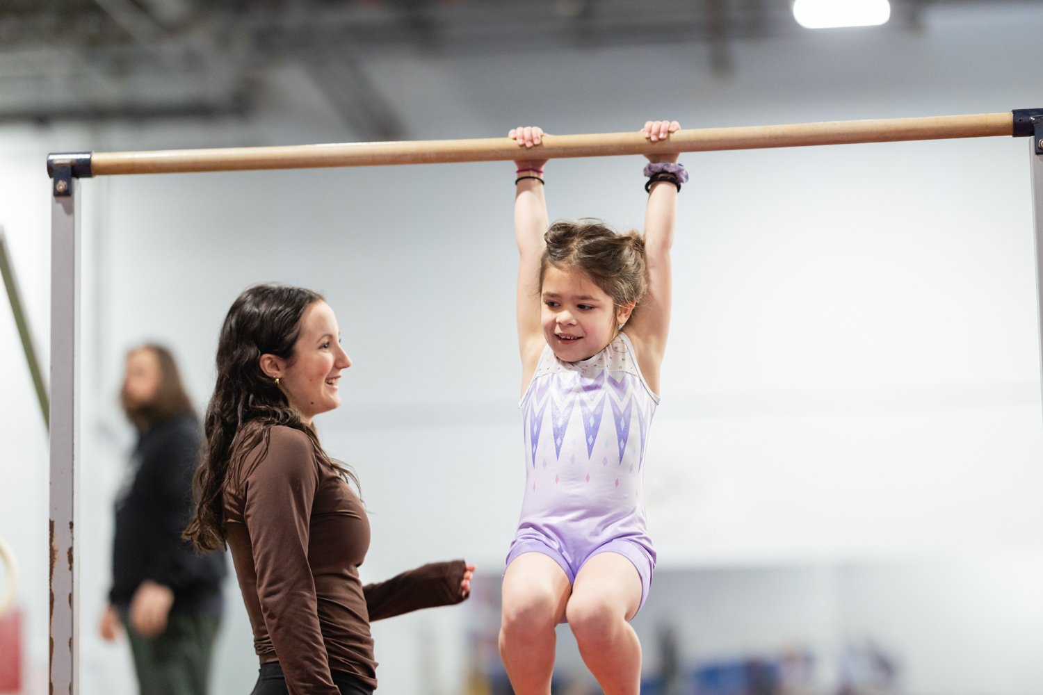 A young girl in a purple gymnastics outfit hanging from a horizontal bar during a practice or class in a gym, with a woman smiling and watching her.