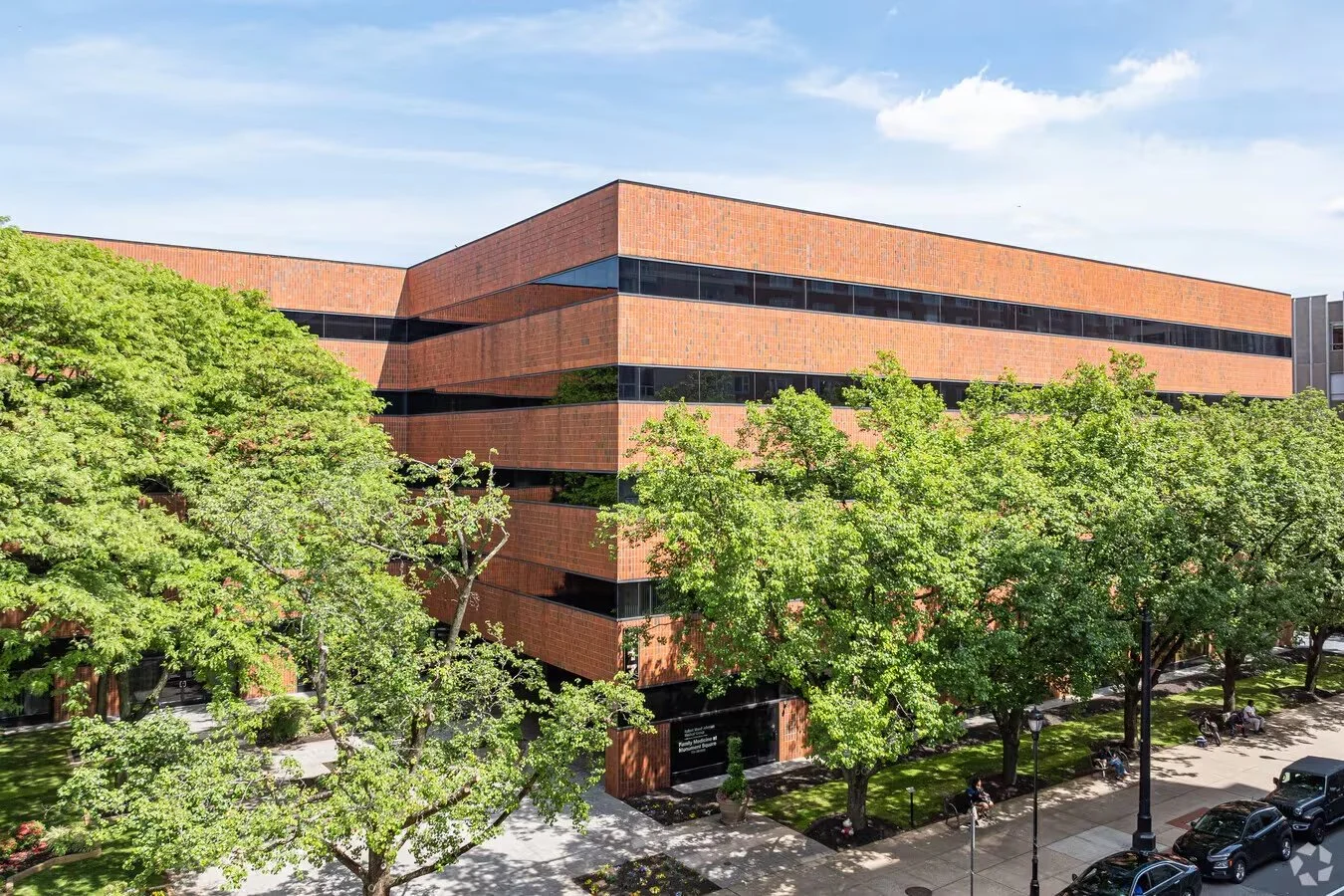 A large, modern brick building with black-tinted windows and green trees in front, located on a city street with parked cars and a few pedestrians.