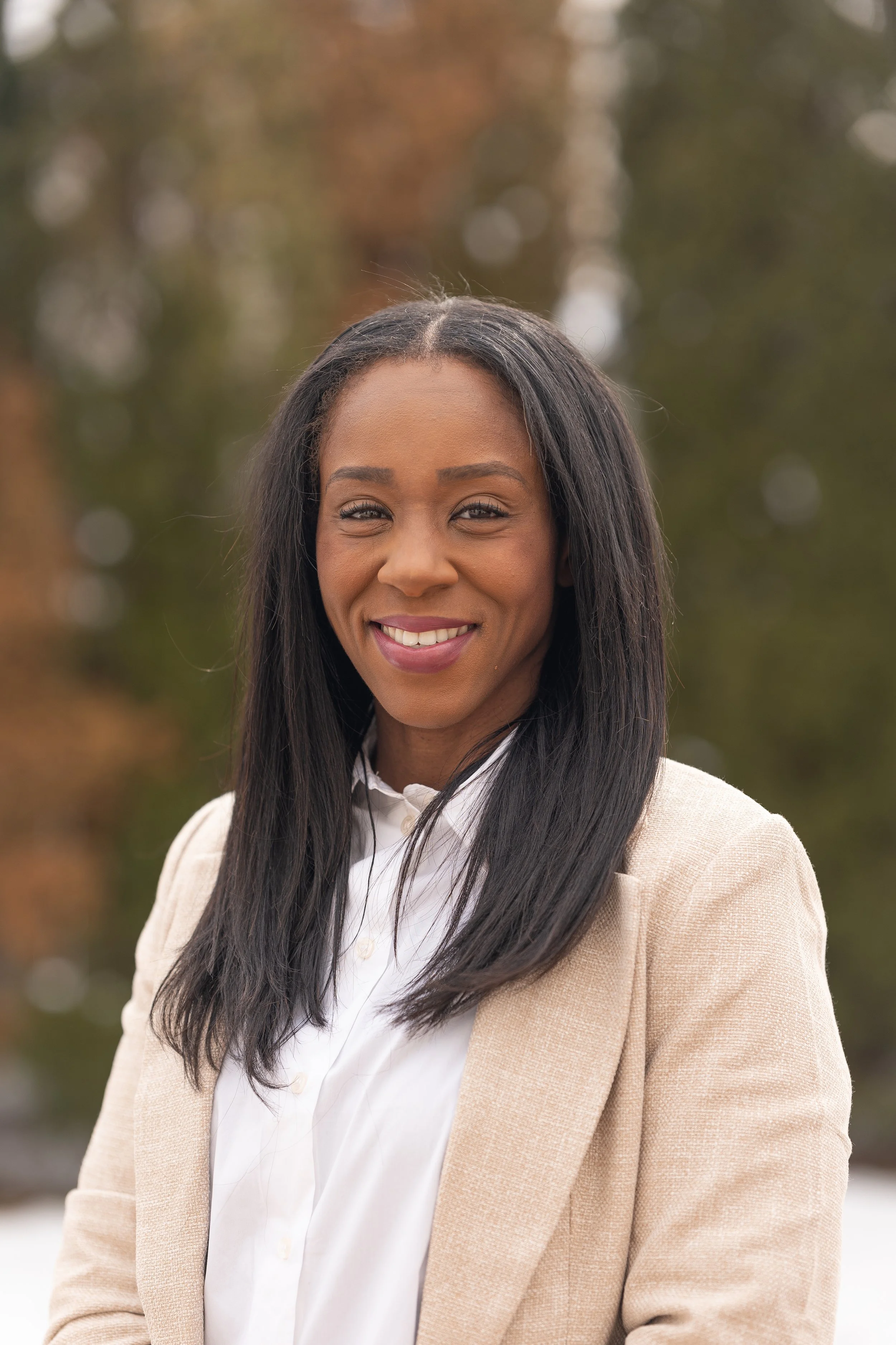 A woman with long black hair smiling, wearing a beige blazer and white shirt, outdoors with blurred trees in the background.
